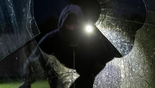 Durch eine eingeschlagene Scheibe blickt ein fiktiver Einbrecher